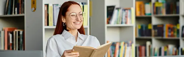 A redheaded woman engrossed in a book, surrounded by tall shelves in a library, absorbing knowledge. — Stock Photo