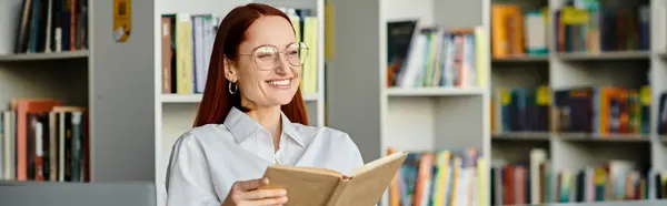 Una donna dai capelli rossi avvolta in un libro, circondata da alti scaffali in una biblioteca, assorbe la conoscenza. — Foto stock