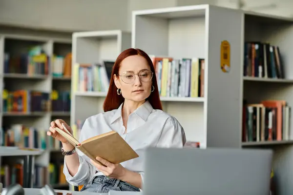 A redheaded woman is deeply engrossed in reading a book inside a library, surrounded by shelves of books. — Stock Photo