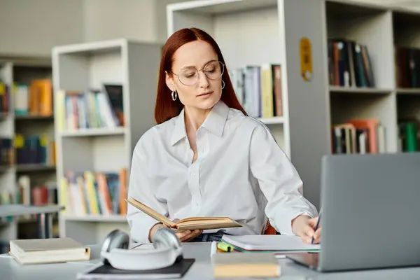 Una mujer pelirroja, una tutora, participa en una sesión de enseñanza en línea usando una computadora portátil en un entorno tranquilo de biblioteca. - foto de stock