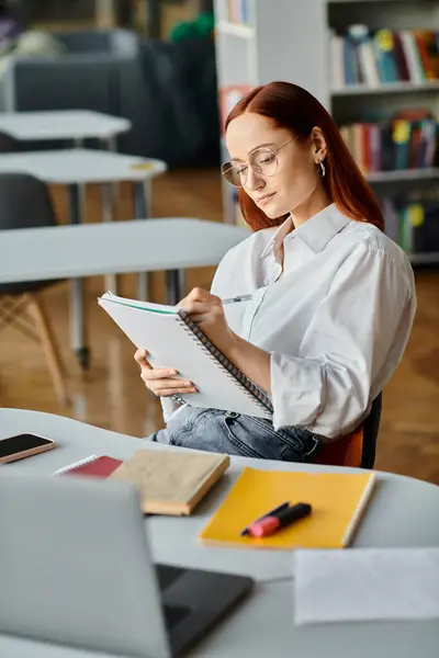 Eine rothaarige Lehrerin sitzt an ihrem Schreibtisch und konzentriert sich darauf, nach einer Online-Lektion in ein Notizbuch zu schreiben.. — Stockfoto