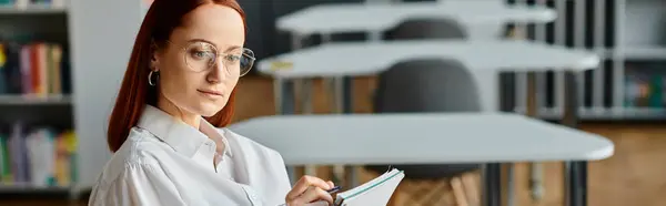 A female tutor with red hair sits in a classroom, diligently teaching online using a laptop after school hours. — Stock Photo