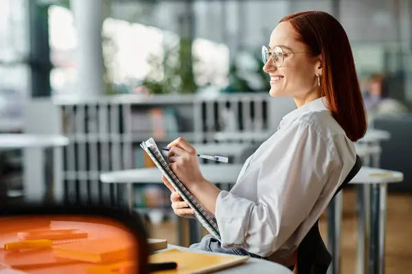 A red-haired woman sits at a desk, writing on a clipboard while teaching an online lesson using a laptop after school. — Stock Photo