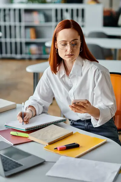 A redhead female tutor providing online teaching services, managing her laptop and cell phone after a school lesson. — Stock Photo