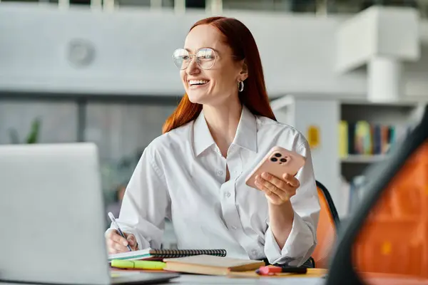 Eine rothaarige Lehrerin unterrichtet nach der Schule online und sitzt mit Laptop und Handy am Schreibtisch. — Stockfoto