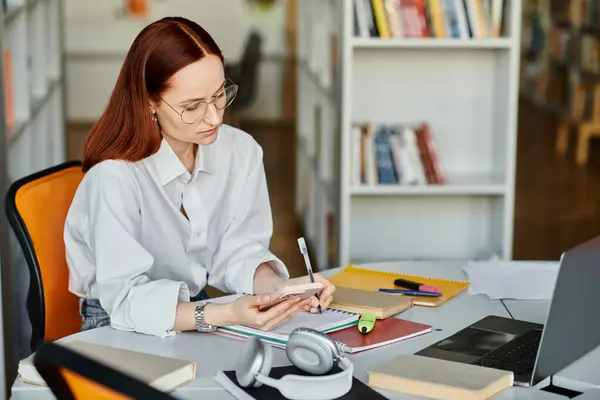 Eine rothaarige Tutorin sitzt an einem Schreibtisch und unterrichtet online mit Laptop und Handy. — Stockfoto