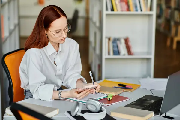 A redhead female tutor sitting at a desk, teaching online with a laptop and cell phone. — Stock Photo