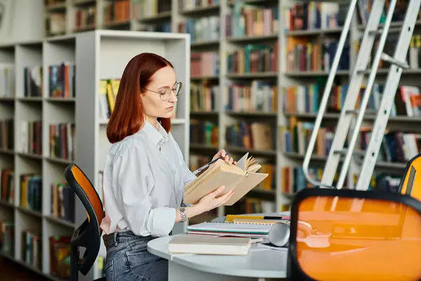 Рыжая женщина сидит за столом в библиотеке, поглощенная чтением книги. — стоковое фото
