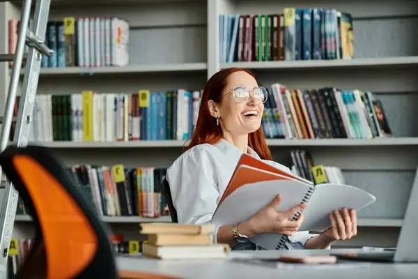 Une tutrice rousse enseigne en ligne, assise à un bureau de bibliothèque absorbé par la lecture d'un livre après sa leçon après l'école. — Photo de stock