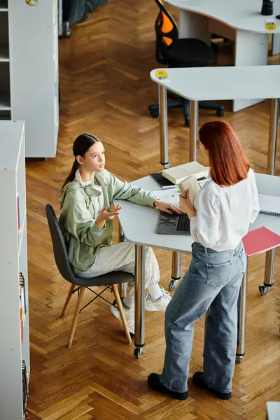 Redhead woman tutors a teenage girl, their study session focused on a laptop in a quiet library setting. — Stock Photo