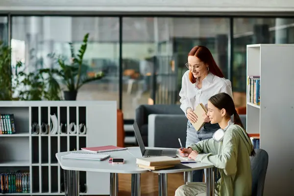 Una donna dai capelli rossi istruisce un'adolescente ad una scrivania in un ufficio moderno, entrambe assorte nei loro studi con un computer portatile. — Foto stock