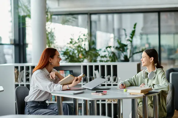 A redhead woman is tutoring a teenage girl at a table in a modern office, using a laptop for after-school lessons. — Stock Photo