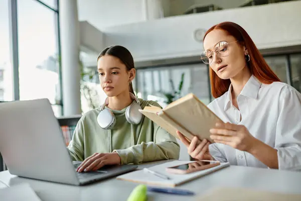 A redhead woman teaches a teenage girl at a table, both immersed in a laptop while reading a book. — Stock Photo