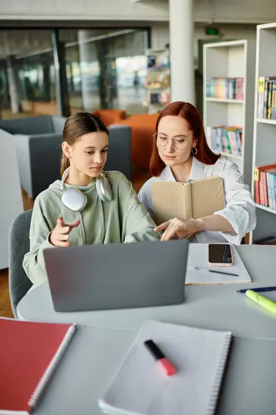 Femme rousse enseignant adolescente, tuteur et élève ayant des leçons après l'école tout en travaillant sur un ordinateur portable dans une bibliothèque. — Photo de stock