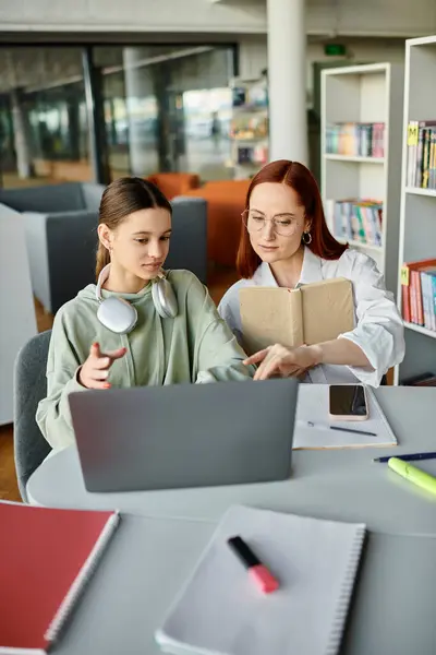 Femme rousse enseignant adolescente, tuteur et élève ayant des leçons après l'école tout en travaillant sur un ordinateur portable dans une bibliothèque. — Photo de stock