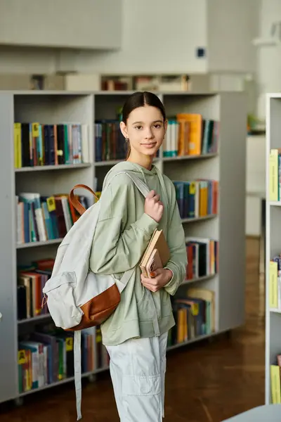 Una chica con una mochila está en una biblioteca, absorta en sus alrededores mientras explora los estantes de los libros. - foto de stock