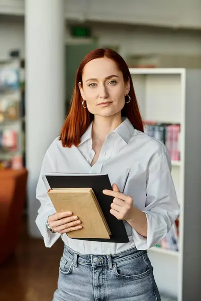 A redheaded woman holds a book in a library, embodying modern education. — Stock Photo