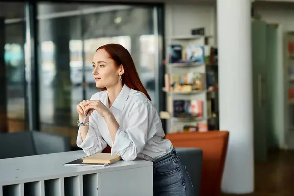 A redhead woman sitting at desk in a library, modern education. — Stock Photo