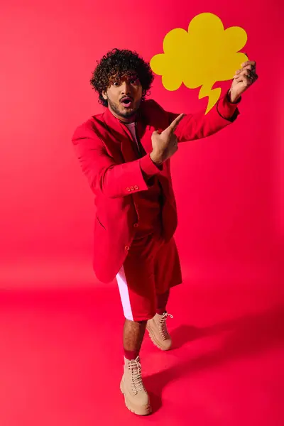 Um jovem indiano em um terno vermelho segurando uma bolha de discurso amarelo em um cenário vívido. — Fotografia de Stock