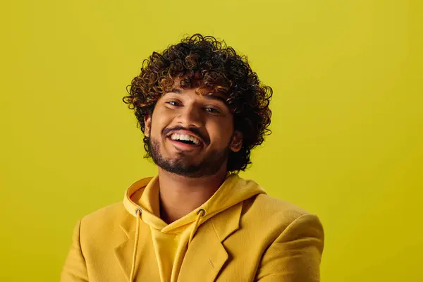 Schöner junger indischer Mann mit lockigem Haar posiert in einem gelben Kapuzenpulli vor einem lebhaften Hintergrund. — Stockfoto