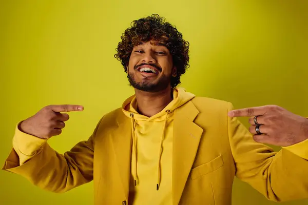 Ein gutaussehender junger indischer Mann in einer gelben Jacke zeigt auf etwas vor einer lebhaften Kulisse. — Stockfoto