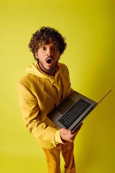 Schöner junger indischer Mann mit lockigem Haar hält einen Laptop vor lebendigem Hintergrund. — Stockfoto