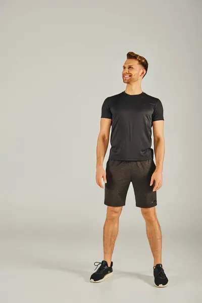 A young athletic man wearing a black t-shirt and shorts, working out in a studio against a grey background. — Stock Photo