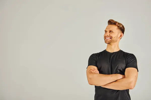 Um jovem atlético em uma camiseta preta posa com os braços cruzados em um estúdio em um fundo cinza. — Fotografia de Stock