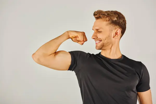 Um jovem atlético em desgaste ativo flexiona seu bíceps com confiança contra um fundo cinza neutro. — Fotografia de Stock