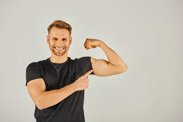 A young athletic man in active wear flexing his biceps against a gray background, showcasing strength and determination. — Stock Photo