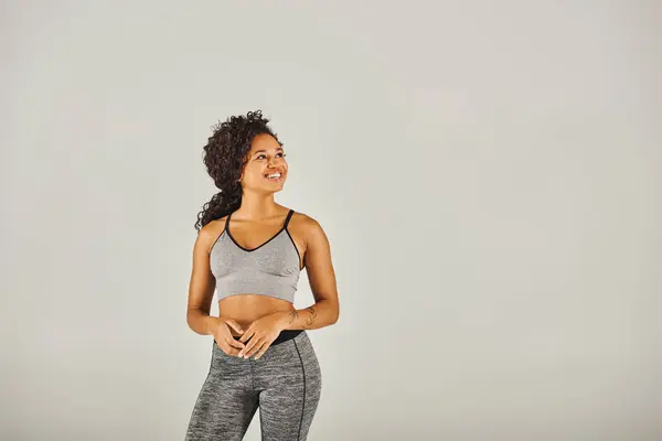 Uma jovem afro-americana em sutiã esportivo e leggings fazendo uma rotina de exercícios em um estúdio contra um fundo cinza. — Fotografia de Stock