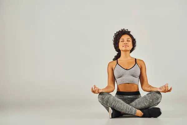 A young African American woman in activewear meditates peacefully on a gray background in a studio setting. — Stock Photo