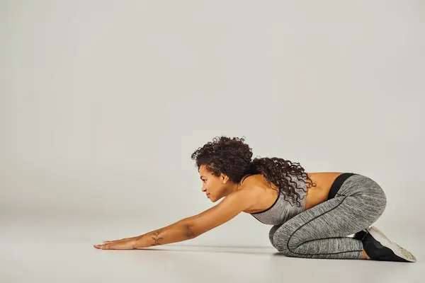 Молодая афроамериканка в активной одежде изящно исполняет позу йоги на безмятежном сером фоне в студии. — стоковое фото