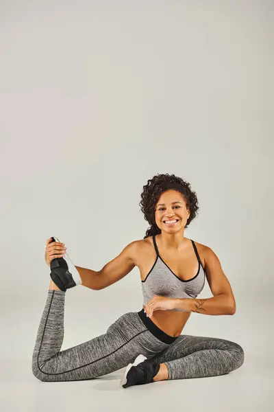 Молодая, стройная афроамериканка в активной одежде практикует позу йоги на сером фоне в спокойной обстановке студии. — стоковое фото