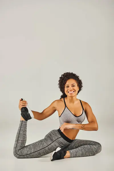 A young, fit African American woman in active wear practices a yoga pose on a grey background in a serene studio setting. — Stock Photo