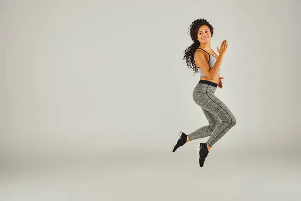 A young African American woman in athletic attire joyfully jumps against a plain gray backdrop in a studio setting. — Stock Photo