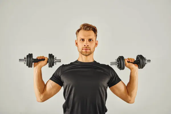 A young sportsman in active wear holds two dumbbells in a studio against a grey background, focusing on fitness and strength. — Stock Photo