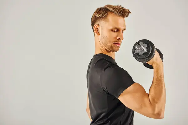 A young sportsman in active wear lifts a dumbbell in a studio with a grey background. — Stock Photo