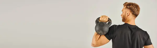 A young sportsman in active wear lifts a kettlebell with determination in a studio setting with a grey background. — Stock Photo