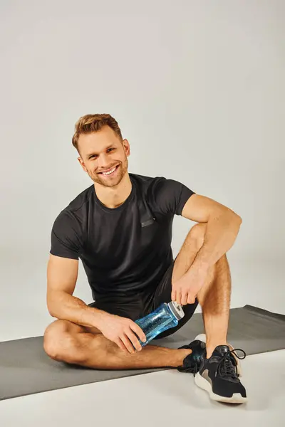Ein junger Sportler in aktiver Kleidung sitzt auf einer Yogamatte, hält eine Wasserflasche in der Hand und macht nach einer Sitzung eine Pause.. — Stockfoto