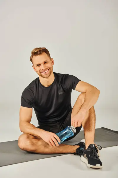 A young sportsman in active wear sits on a yoga mat, holding a water bottle, taking a break after a session. — Stock Photo
