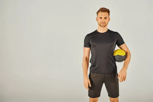 Jovem desportista de t-shirt preta demonstrando exercício físico com uma bola amarela e preta vibrante em estúdio. — Fotografia de Stock