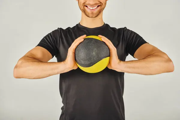 Jeune sportif en tenue active tenant énergiquement une boule jaune et noire vibrante dans un studio au fond gris. — Photo de stock