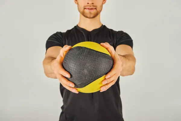 A young sportsman in active wear holds a yellow and black ball in a studio with a grey background. — Stock Photo