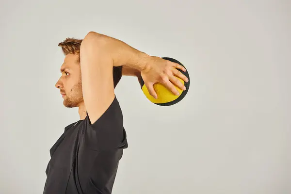 Молодий спортсмен в активному носінні енергійно вправляється з жовтим м'ячем в студії з сірим фоном. — стокове фото