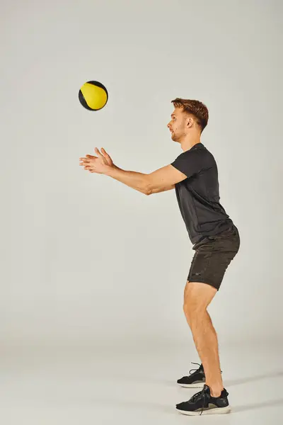 A young sportsman in active wear catches a yellow and black ball in a dynamic studio setting with a grey background. — Stock Photo