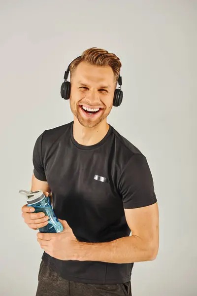 A young sportsman in active wear, wearing headphones and holding a water bottle in a studio with a grey background. — Stock Photo