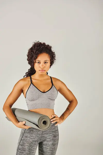 Кучерява афроамериканська спортсменка в активному одязі тримає йога килимок в студії з сірим фоном. — стокове фото
