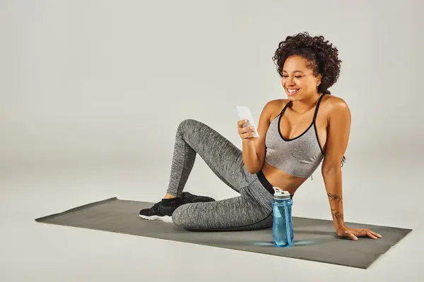 Une sportive afro-américaine frisée en tenue active assise sur un tapis de yoga, naviguant sur son téléphone, sur un fond gris. — Photo de stock