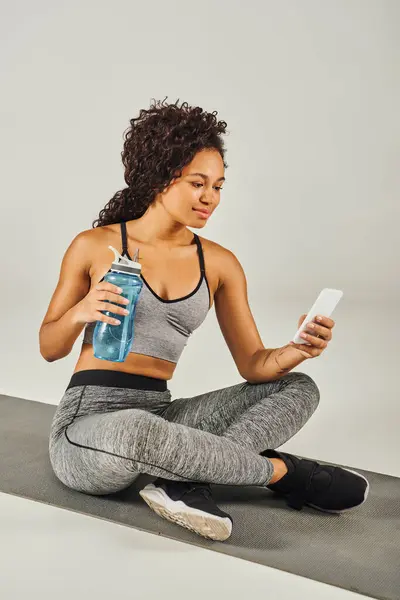 A curly African American sportswoman is sitting on a yoga mat, holding a water bottle in a yoga studio with a grey background. — Stock Photo